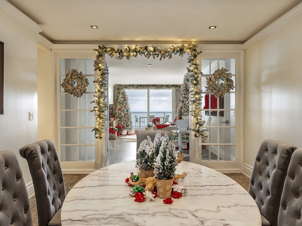 Festively decorated coastal dining room at Pelican Grand Beach Resort in Fort Lauderdale, featuring a marble table, tufted gray chairs, holiday garland-draped French doors, and an oceanview Christmas tree.