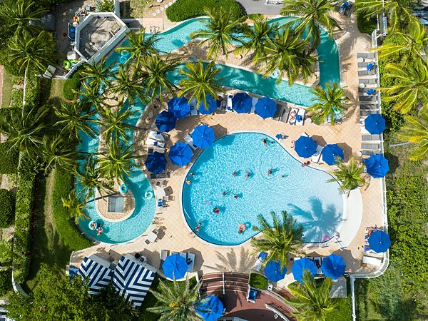 Aerial view of the zero-entry pool and winding lazy river surrounded by palm trees and blue umbrellas at Pelican Grand Beach Resort in Fort Lauderdale, Florida