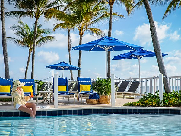 Woman in sun hat relaxing poolside with a tropical cocktail under blue umbrellas at Pelican Grand Beach Resort in Fort Lauderdale, Florida